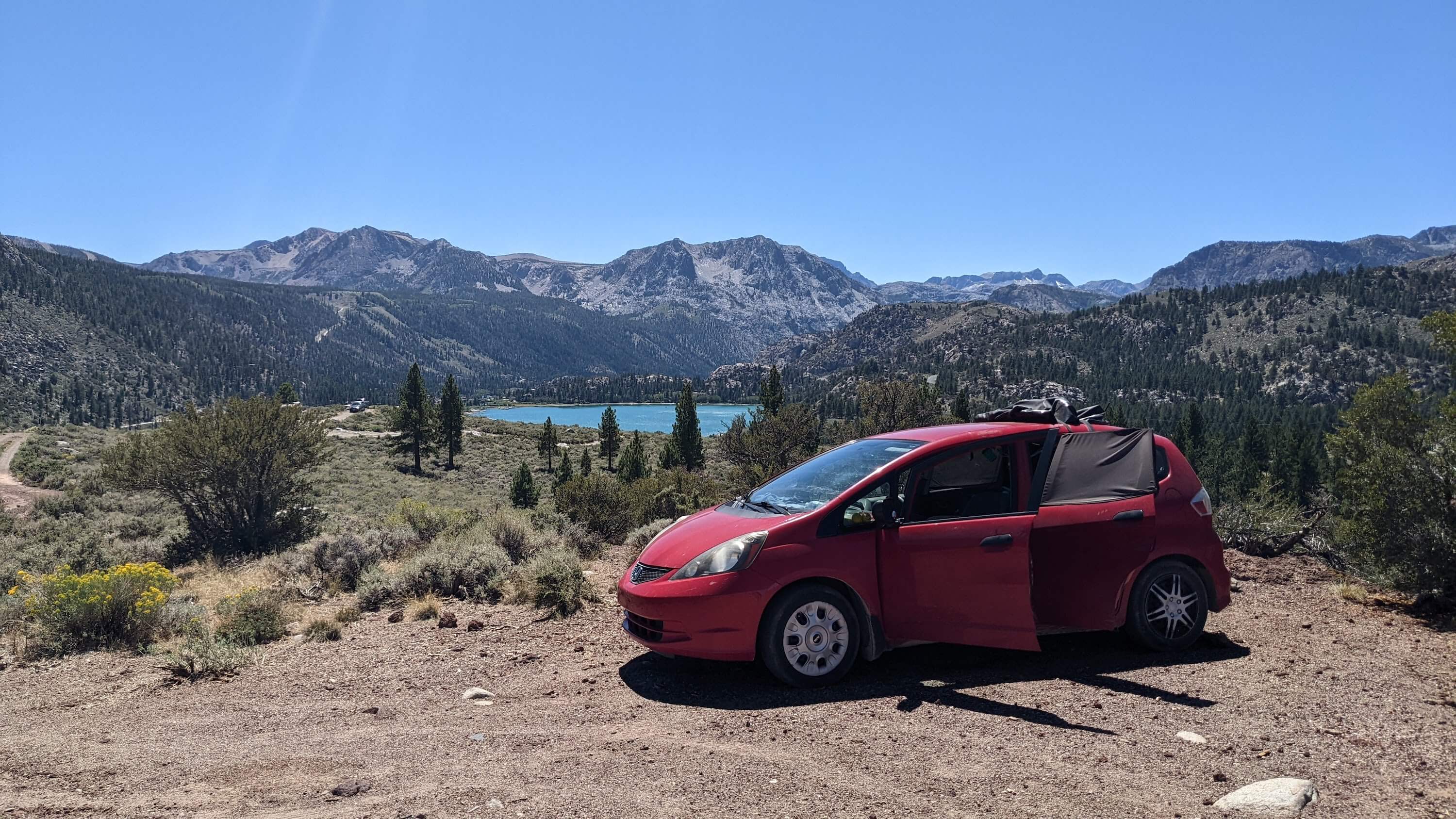 Red Honda Fit 2013 at the top of a mountain overlooking mountains and a lake on a bright sunny day